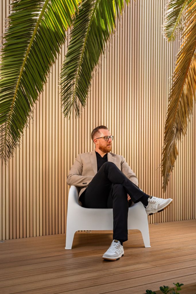 Man in a beige blazer seated on a timber deck under palm fronds